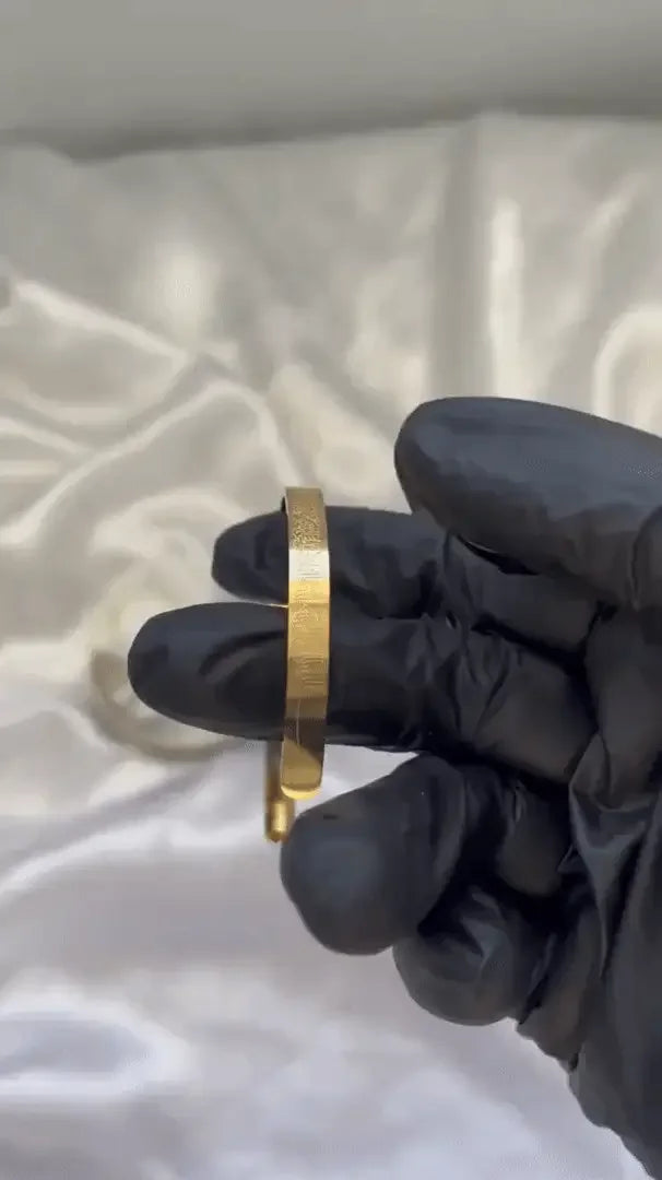 A person wearing black gloves holding a gold-plated engraved cuff bracelet.