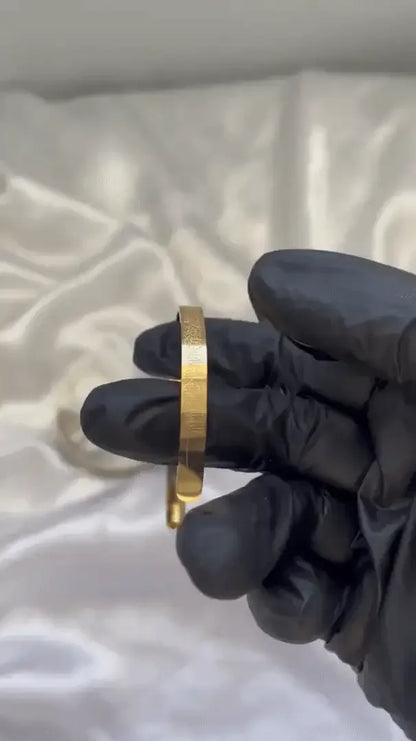 A person wearing black gloves holding a gold-plated engraved cuff bracelet.